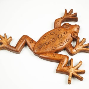 May include: A brown wooden carving of a frog with a smooth, polished finish. The figure features incised circular patterns along its back and limbs, with four splayed feet and a defined head, presented in a flat, stylised, and symmetrical form.