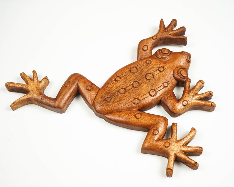 May include: A brown wooden carving of a frog with a smooth, polished finish. The figure features incised circular patterns along its back and limbs, with four splayed feet and a defined head, presented in a flat, stylised, and symmetrical form.