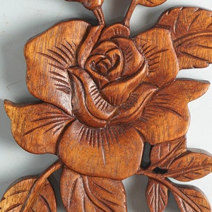 K&ouml;nnte beinhalten: Eine braune Holzwandschnitzerei, die eine detaillierte Rose mit geschichteten Bl&uuml;tenbl&auml;ttern und mehreren Bl&auml;ttern darstellt. Die Holzmaserung ist auf der gesamten Oberfl&auml;che sichtbar und die Schnitzerei zeigt tiefe, texturierte Linien.