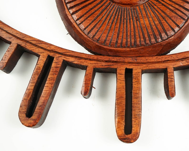 May include: A brown wooden wall decor piece featuring a central circular element with carved radial lines, surrounded by a series of downward-pointing, elongated rectangular cutouts that resemble eyelashes or rays in a sunburst pattern.