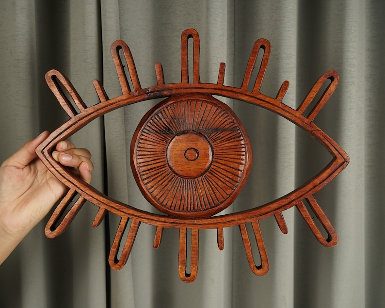 May include: A large, hand-carved wooden wall hanging shaped like an eye. The piece features a central circular iris with radiating lines and an outer almond-shaped frame with elongated, finger-like projections representing eyelashes in a warm brown tone.