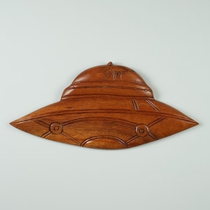 Peut inclure: Une sculpture en bois d'une soucoupe volante, avec une finition en bois brun. La soucoupe a un dessus arrondi et un fond plat, avec des d&eacute;tails sculpt&eacute;s dans la surface.
