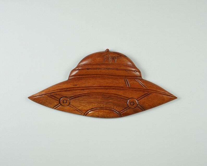 Peut inclure: Une sculpture en bois d'une soucoupe volante, avec une finition en bois brun. La soucoupe a un dessus arrondi et un fond plat, avec des d&eacute;tails sculpt&eacute;s dans la surface.