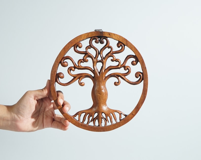 May include: A round wooden wall hanging with a carved tree of life design. The tree has intricate branches and roots, and the wood is a rich brown color.