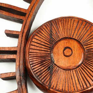 May include: A dark brown wooden wall decor piece featuring a circular center with carved radial lines and a small central circle. The surrounding frame has protruding rectangular segments that resemble eyelashes or sun rays, crafted from wood.
