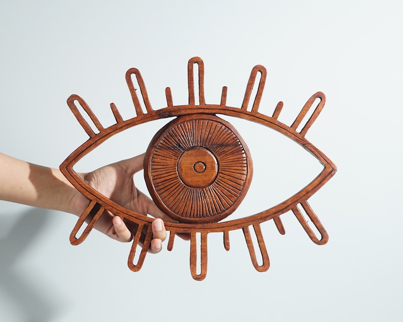May include: A brown wooden wall hanging shaped like an eye with a central circular iris featuring carved radial lines and an outer almond-shaped frame with elongated vertical slats representing eyelashes extending from the top and bottom edges.