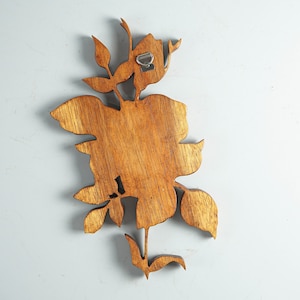K&ouml;nnte beinhalten: Ein brauner Wandbehang aus Holz in Form einer Blume mit Bl&auml;ttern und einem Stiel. Das St&uuml;ck weist eine sichtbare Holzmaserung auf und verf&uuml;gt &uuml;ber eine silberne Metallhalterung an der oberen R&uuml;ckseite zur Befestigung an einer Wand.