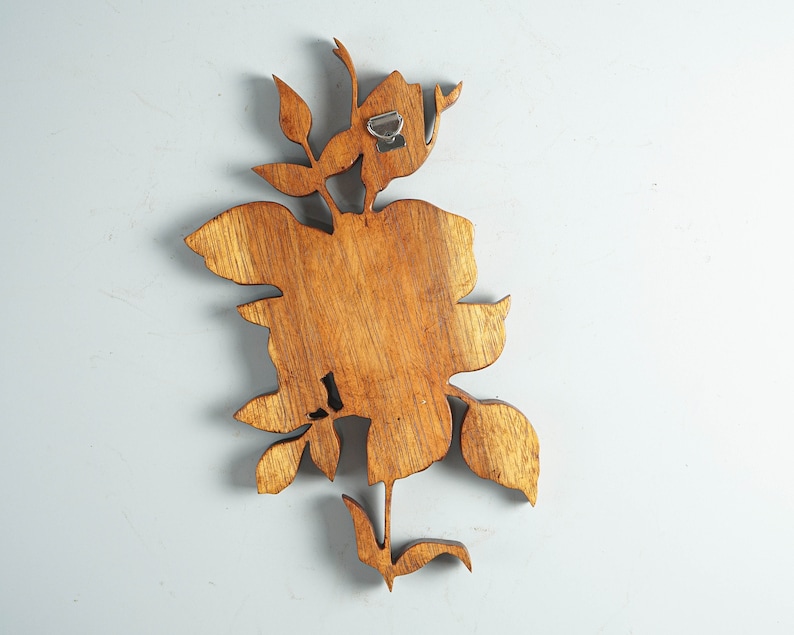 K&ouml;nnte beinhalten: Ein brauner Wandbehang aus Holz in Form einer Blume mit Bl&auml;ttern und einem Stiel. Das St&uuml;ck weist eine sichtbare Holzmaserung auf und verf&uuml;gt &uuml;ber eine silberne Metallhalterung an der oberen R&uuml;ckseite zur Befestigung an einer Wand.