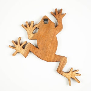 May include: A flat, brown wooden cutout shaped like a frog in a leaping position. The wood grain is visible across the surface. A small metal D-ring hanger is attached to the top centre of the back, allowing the object to be mounted on a wall.