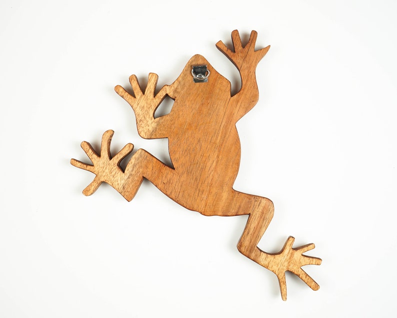 May include: A flat, brown wooden cutout shaped like a frog in a leaping position. The wood grain is visible across the surface. A small metal D-ring hanger is attached to the top centre of the back, allowing the object to be mounted on a wall.