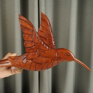 May include: A hand-carved wooden hummingbird sculpture in a reddish-brown color. The hummingbird is in flight with its wings spread out and its beak pointed forward.