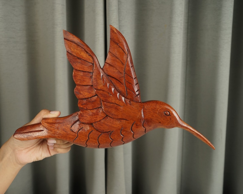 May include: A hand-carved wooden hummingbird sculpture in a reddish-brown color. The hummingbird is in flight with its wings spread out and its beak pointed forward.