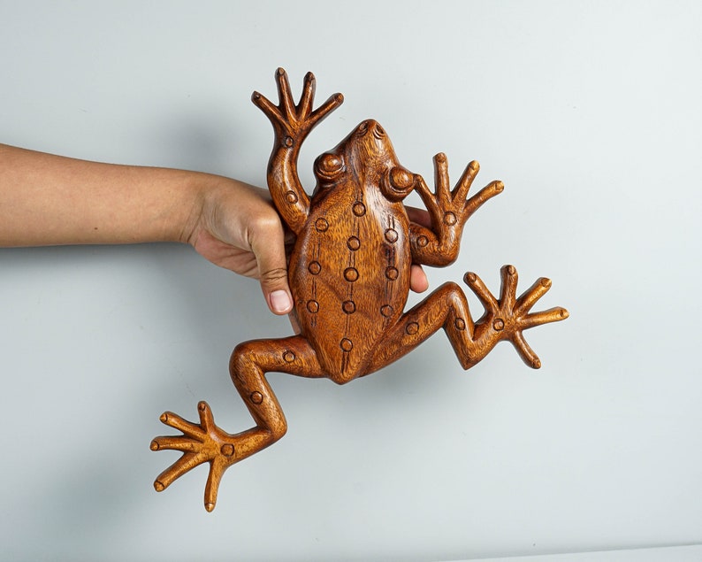 May include: A carved wooden frog figurine with a smooth, polished brown finish. The surface features a series of small, circular indentations arranged in vertical lines along the back and legs, with detailed toes on each of the four splayed limbs.