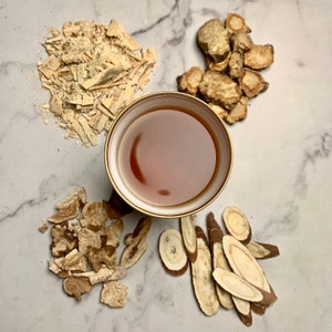 May include: A white ceramic bowl with a gold rim filled with a dark brown liquid.  Surrounding the bowl are four piles of dried herbs, including light brown, dark brown, and tan pieces.