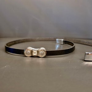 May include: A black leather bracelet with a silver bike chain link clasp.