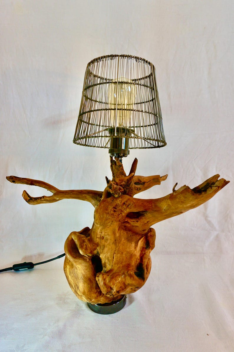Puede incluir: Una l&aacute;mpara de mesa con una pantalla de jaula de alambre se asienta sobre una base de madera que se asemeja a un tronco de &aacute;rbol. La l&aacute;mpara est&aacute; encendida y la bombilla es visible a trav&eacute;s de la jaula de alambre.