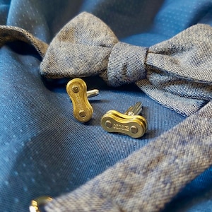 May include: A pair of gold cufflinks made from a bicycle chain link. The cufflinks are on a blue fabric background with a bow tie.