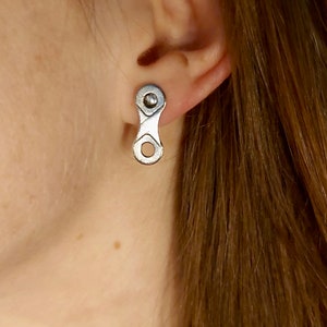 May include: A silver-toned stud earring shaped like a single bicycle chain link. The earring features a smooth, metallic finish with a circular opening at the bottom and a central post attachment point, securely fastened to the earlobe.