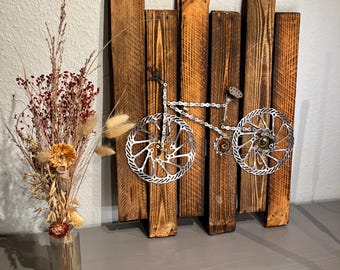 Puede incluir: Una escultura de bicicleta de metal hecha con piezas de bicicleta, montada sobre un fondo de madera rústica. La escultura está pintada en plata y presenta una cadena, engranajes y rotores de freno.