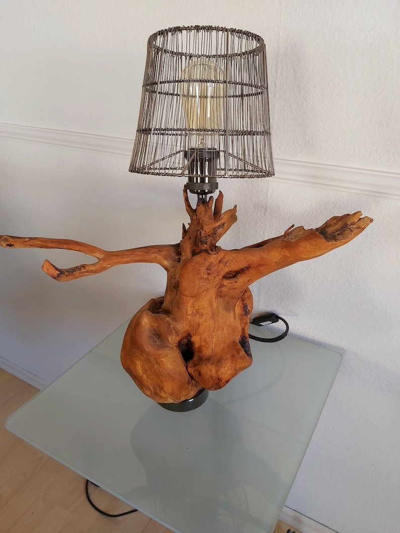 Puede incluir: Una l&aacute;mpara de mesa r&uacute;stica con una pantalla de jaula de alambre y una base de madera con forma de rama de &aacute;rbol. La l&aacute;mpara est&aacute; sobre una mesa de vidrio.