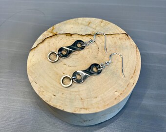 Puede incluir: Un par de pendientes plateados. Cada pendiente presenta un diseño de eslabón de cadena de bicicleta con un pequeño anillo circular. Los pendientes se exhiben sobre una superficie de madera redonda y clara.
