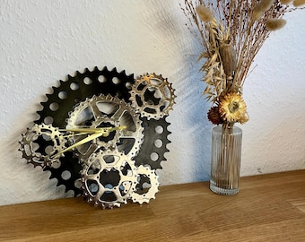 Puede incluir: Un reloj hecho de engranajes de bicicleta plateados y negros, con manecillas doradas, montado en una pared de madera.