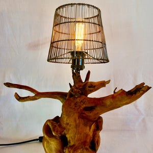 Puede incluir: Una l&aacute;mpara de mesa con una pantalla de jaula de alambre y una base de madera con forma de ra&iacute;z de &aacute;rbol. La l&aacute;mpara est&aacute; encendida y la bombilla es visible.