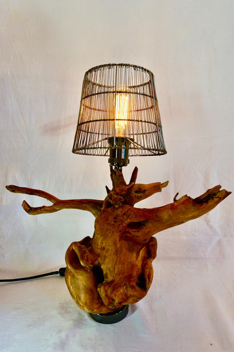 Puede incluir: Una l&aacute;mpara de mesa con una pantalla de jaula de alambre y una base de madera con forma de ra&iacute;z de &aacute;rbol. La l&aacute;mpara est&aacute; encendida y la bombilla es visible.