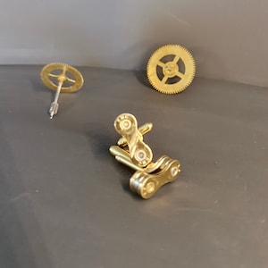 May include: Gold-toned cufflinks made from a bicycle chain link. The cufflinks are displayed on a grey surface with two gold-toned gears.