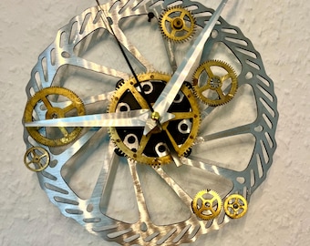 Puede incluir: Un reloj de pared único con un rotor de freno de disco de bicicleta plateado como esfera. El reloj tiene engranajes dorados y manecillas plateadas. El reloj está montado en una pared blanca.