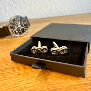 May include: A pair of silver cufflinks in the shape of a bicycle chain link, in a black box with a black velvet interior.