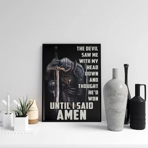 May include: Black framed wall art featuring a knight with a sword, and the white text: "THE DEVIL SAW ME WITH MY HEAD DOWN AND THOUGHT HE'D WON UNTIL I SAID AMEN". Decorative items are in the foreground.