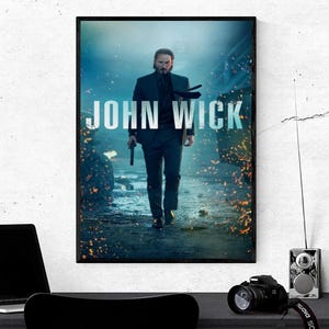 May include: A framed movie poster for "John Wick" featuring a man in a suit holding a gun, walking towards the viewer. The title is in large, distressed white letters. The background is a blurred, action-packed scene.