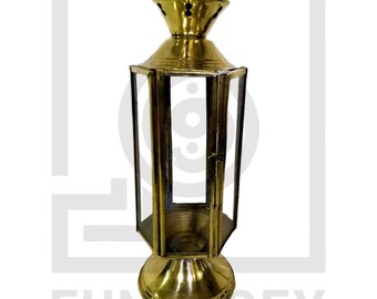 May include: A gold metal lantern with a glass body and a handle on top. The lantern has a decorative design on the top and bottom.