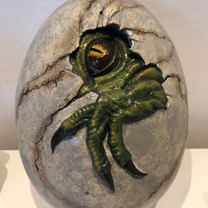 Könnte beinhalten: Eine Keramikskulptur eines Dinosauriers, der aus einem rissigen Ei schlüpft. Der grüne Fuß und das Auge des Dinosauriers sind sichtbar.