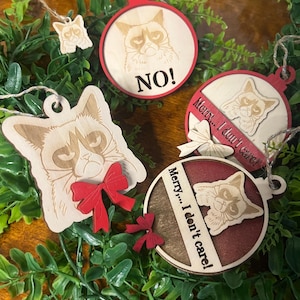 Op de afbeelding: Houten kerstversieringen met een chagrijnig kattenontwerp. De ornamenten hebben verschillende vormen, waaronder een cirkel en een katten silhouet, met rode accenten en de zinnen "NO!" en "Merry... I don't care!"