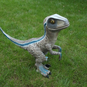 Może przedstawiać: Szara i niebieska zabawka dinozaura z żółtym okiem. Dinozaur stoi na zielonej trawie.
