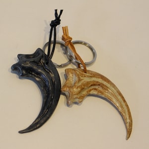 Può includere: Due portachiavi a forma di artiglio di dinosauro, uno nero e uno marrone, attaccati a un portachiavi argentato con un cordino in pelle nera e un cordino in pelle marrone.