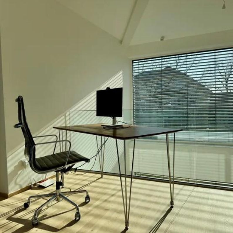 May include: A modern office setup featuring a dark wood desk with metal hairpin legs, a black ergonomic chair, and a computer monitor. The room has a large window with blinds, and the walls are painted a light color.