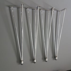 May include: Set of five silver hairpin table legs. Each leg has a flat mounting plate and tapers to a pointed end. The legs are made of shiny metal and are arranged against a gray background. These legs are designed for furniture construction.