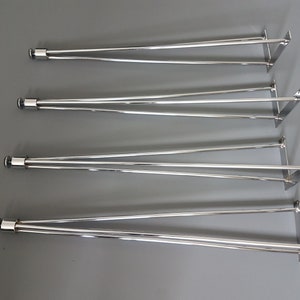 May include: Four sets of silver-colored metal table legs. Each leg set has a cylindrical top with a black cap, two angled support rods, and a square mounting plate. The legs are arranged on a gray surface.