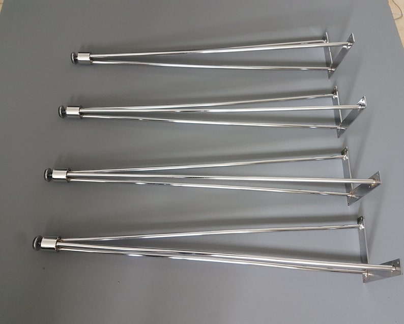 May include: Four sets of silver-colored metal table legs. Each leg set has a cylindrical top with a black cap, two angled support rods, and a square mounting plate. The legs are arranged on a gray surface.