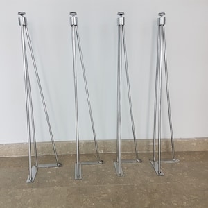 May include: Four silver metal table legs with a modern design. Each leg has a rectangular base, a central support, and three angled supports. The legs are topped with a small, round, silver cap. The legs are designed for furniture.