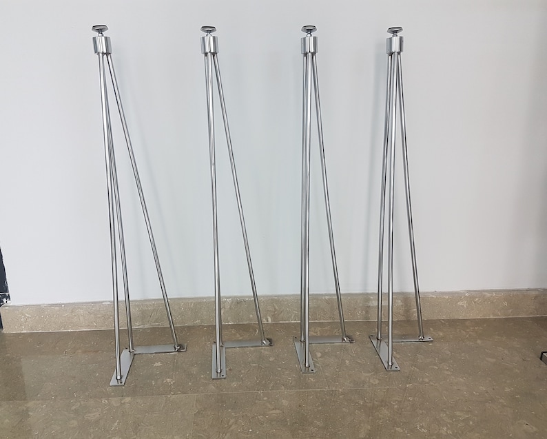 May include: Four silver metal table legs with a modern design. Each leg has a rectangular base, a central support, and three angled supports. The legs are topped with a small, round, silver cap. The legs are designed for furniture.