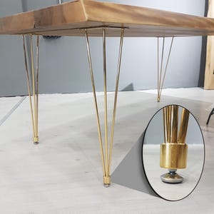 May include: A wooden table with a natural finish and gold-colored hairpin legs. The table's top is rectangular and has a live edge. The legs are thin and angled, with a cylindrical base. The table is approximately 30 inches high.