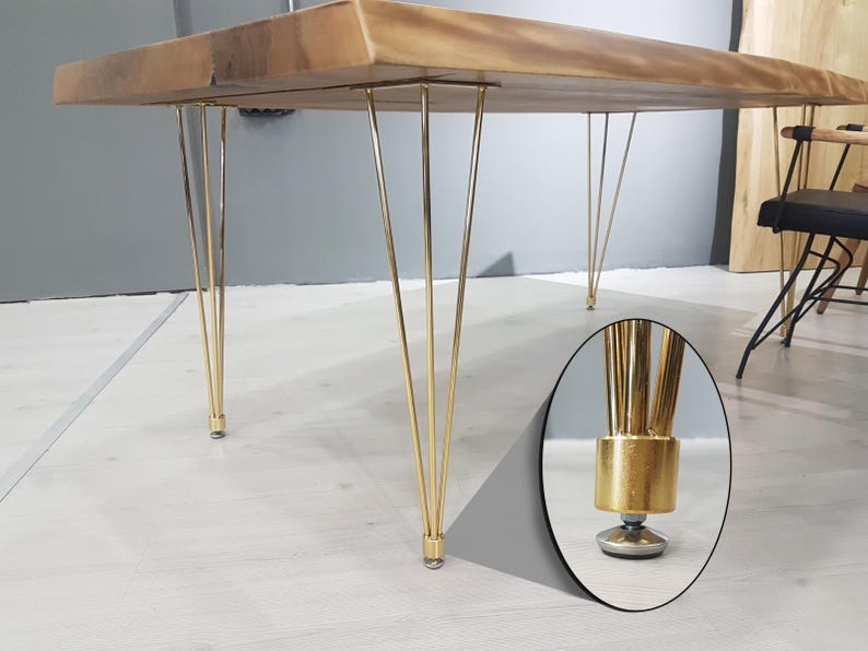 May include: A wooden table with a natural finish and gold-colored hairpin legs. The table's top is rectangular and has a live edge. The legs are thin and angled, with a cylindrical base. The table is approximately 30 inches high.