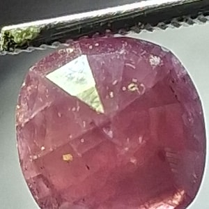 May include: A cushion-cut gemstone with a deep pinkish-red hue and a faceted surface. The stone displays internal inclusions and a distinct triangular light reflection on its top facet, held securely by a pair of metal tweezers.