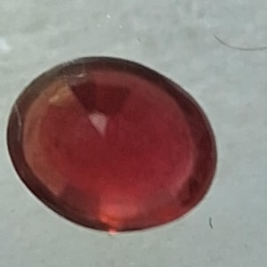 May include: A deep red, oval-shaped gemstone with a smooth, polished surface. The stone exhibits a translucent quality with subtle internal light reflections and a consistent, rich crimson hue throughout its rounded, symmetrical form.