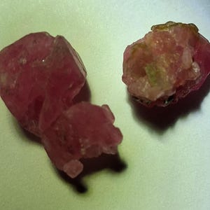 May include: Two raw, unpolished mineral specimens with a deep pink and magenta coloration. The stones feature irregular, jagged edges and a crystalline, translucent texture with visible internal fractures and small, lighter-colored inclusions.