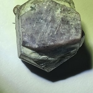 May include: A raw, hexagonal crystal specimen with a muted purple and grey coloration. The stone features a flat, six-sided top surface with visible natural striations and rough, textured edges along the sides of the mineral formation.